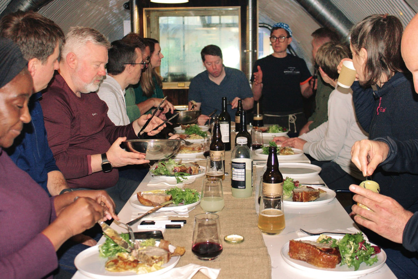 A group of Smoke School workshop attendees enjoying a hearty roast pork lunch at a long table.