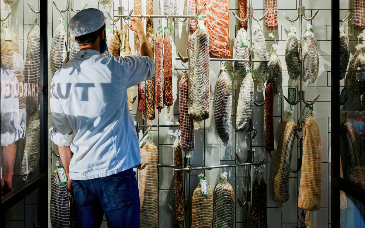 London Smoke & Cure's head of production stands in front of a tiled chamber, inspecting cured meats hanging on racks.