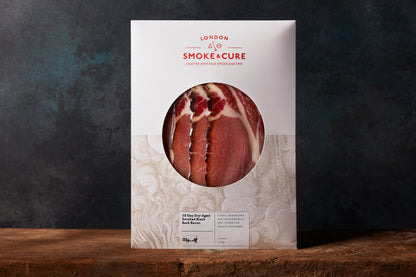 A pack of London Smoke & Cure's Aged Smoked Black Back Bacon standing on a wooden table with a dark background.