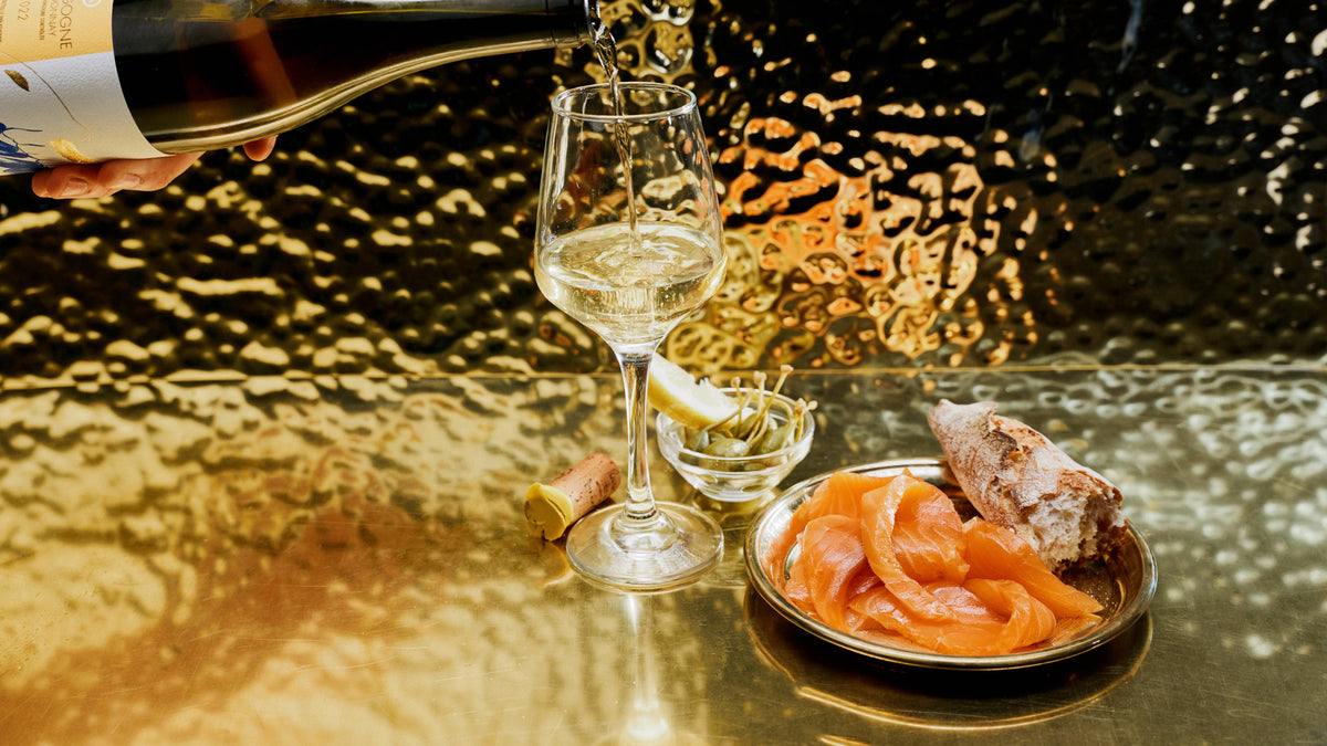 White wine is poured from a bottle into a wine glass on a reflective gold-toned table, with a small plate of sliced cured salmon and a piece of crusty bread nearby.